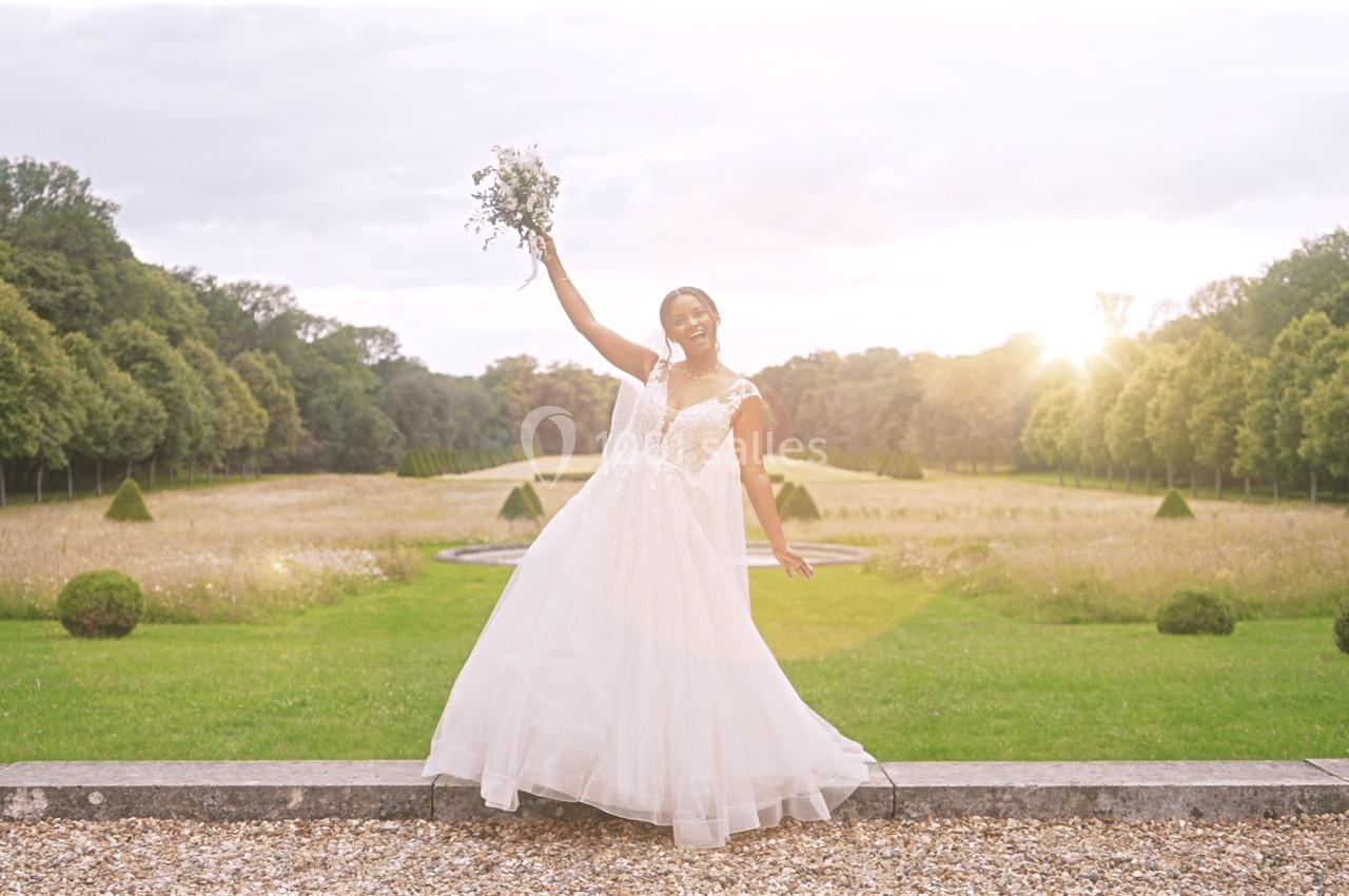 Femme en robe de mariée blanche tenant un bouquet, debout dans un jardin avec une allée bordée d'arbres au coucher du soleil.
