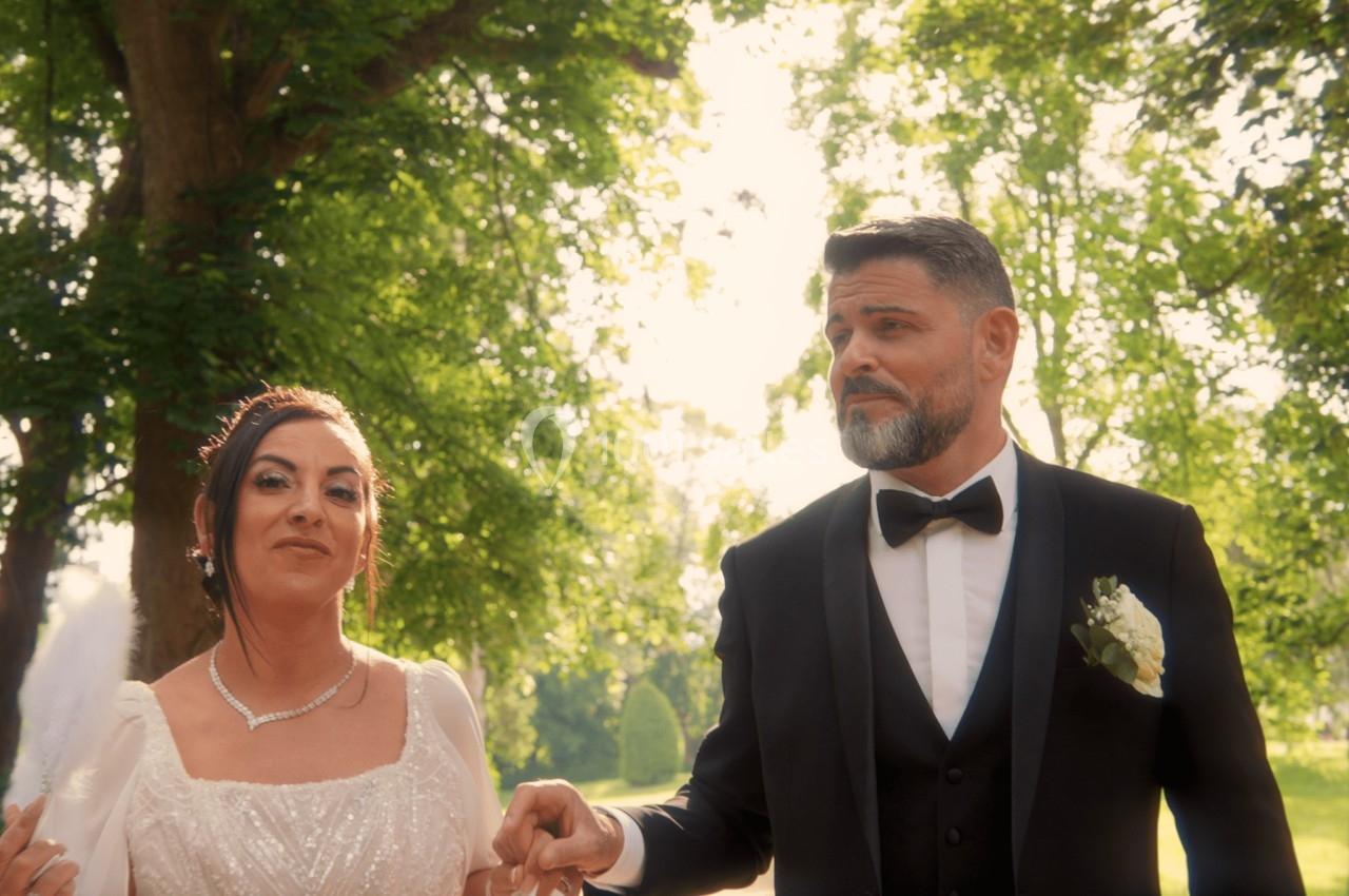 Un couple en tenue de mariage marche dans un jardin verdoyant sous une lumière naturelle.
