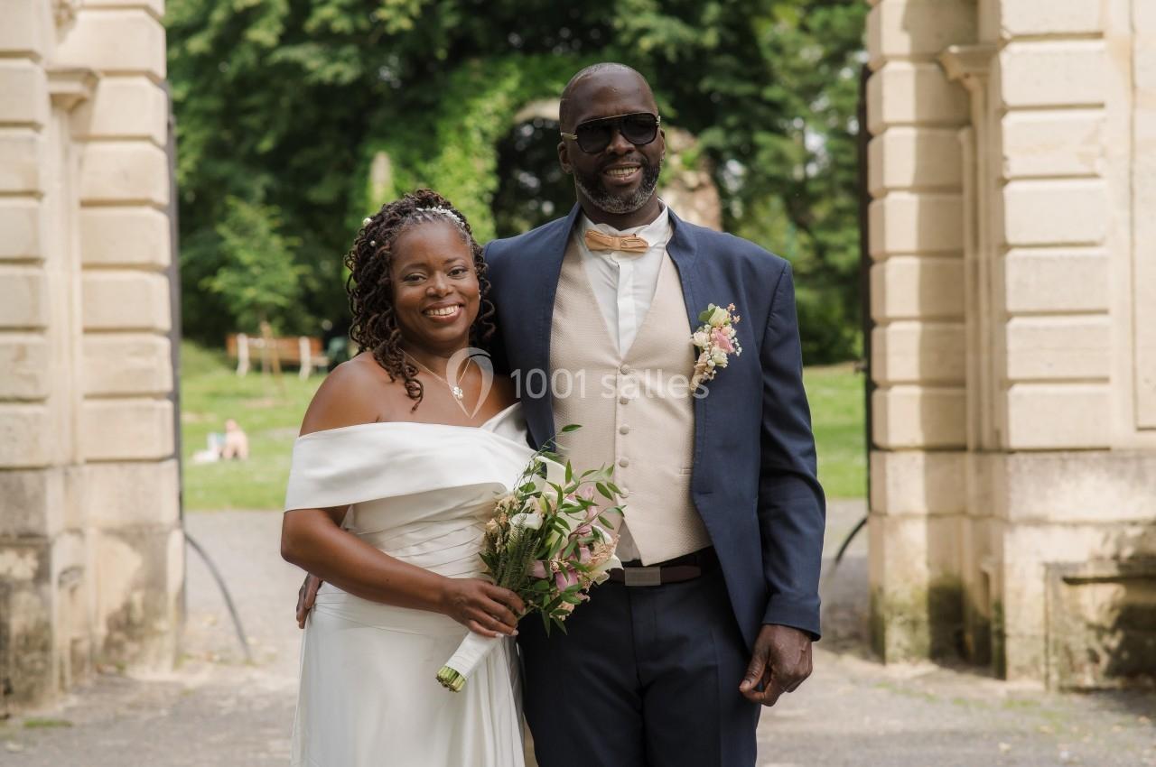 Un couple habillé en tenue de mariage pose devant une arche en pierre dans un parc verdoyant.