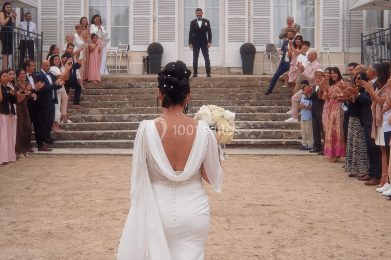Une mariée avance vers un marié debout en haut d'escaliers, entourés d'invités applaudissant.