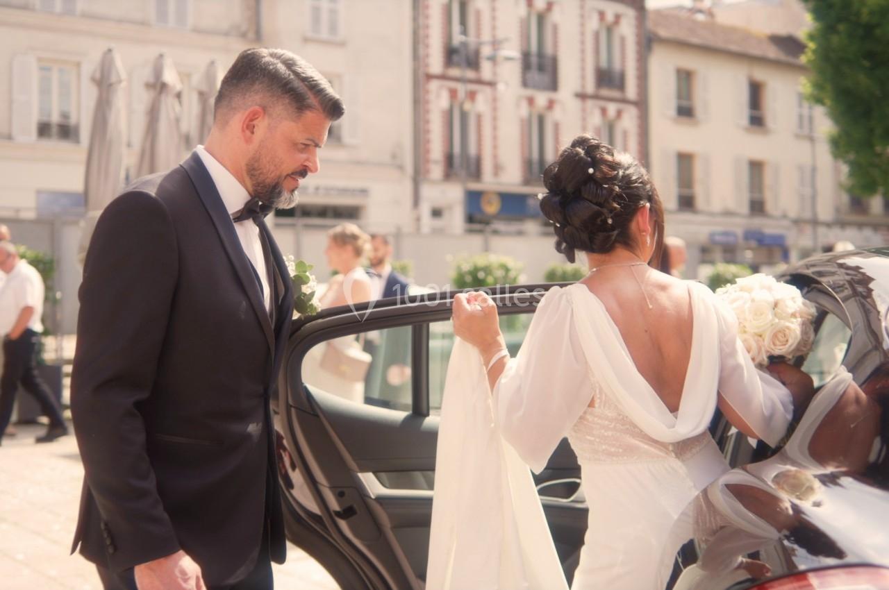 Un homme en costume observe une femme en robe de mariée entrant dans une voiture devant des bâtiments urbains.