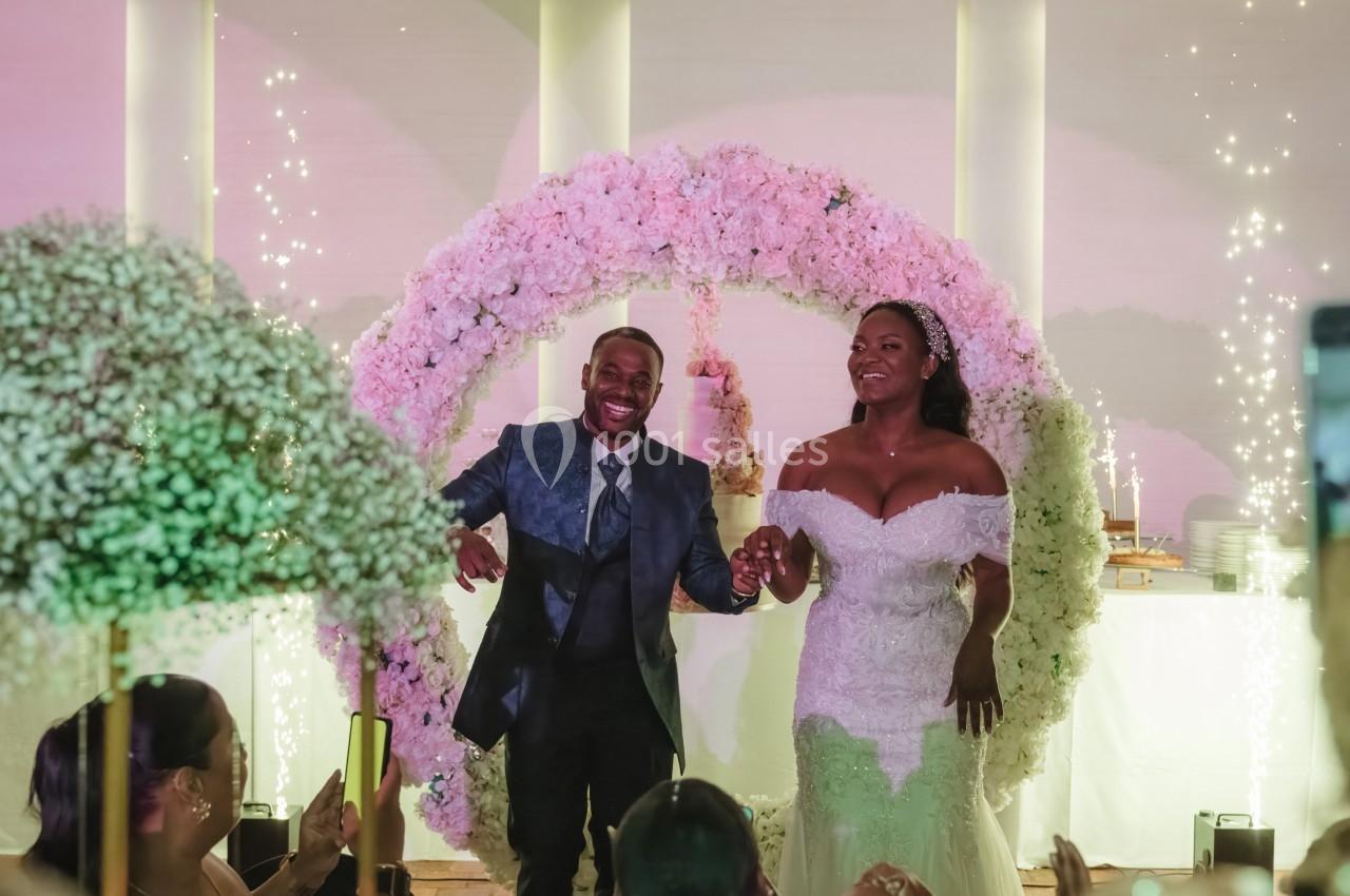 Un couple souriant en tenue de mariage devant une arche décorée de fleurs roses et des étincelles en arrière-plan.