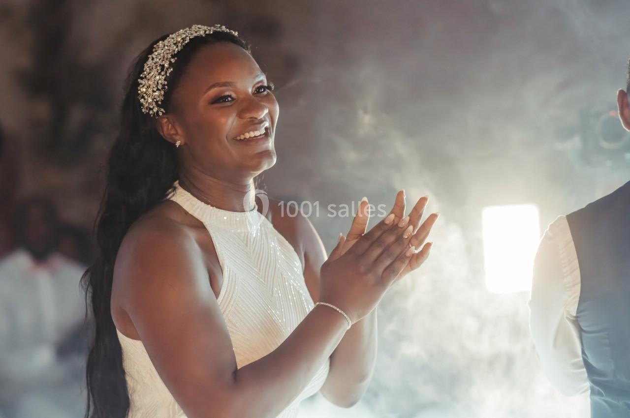 Femme souriante en robe blanche applaudissant dans un environnement lumineux et légèrement enfumé.