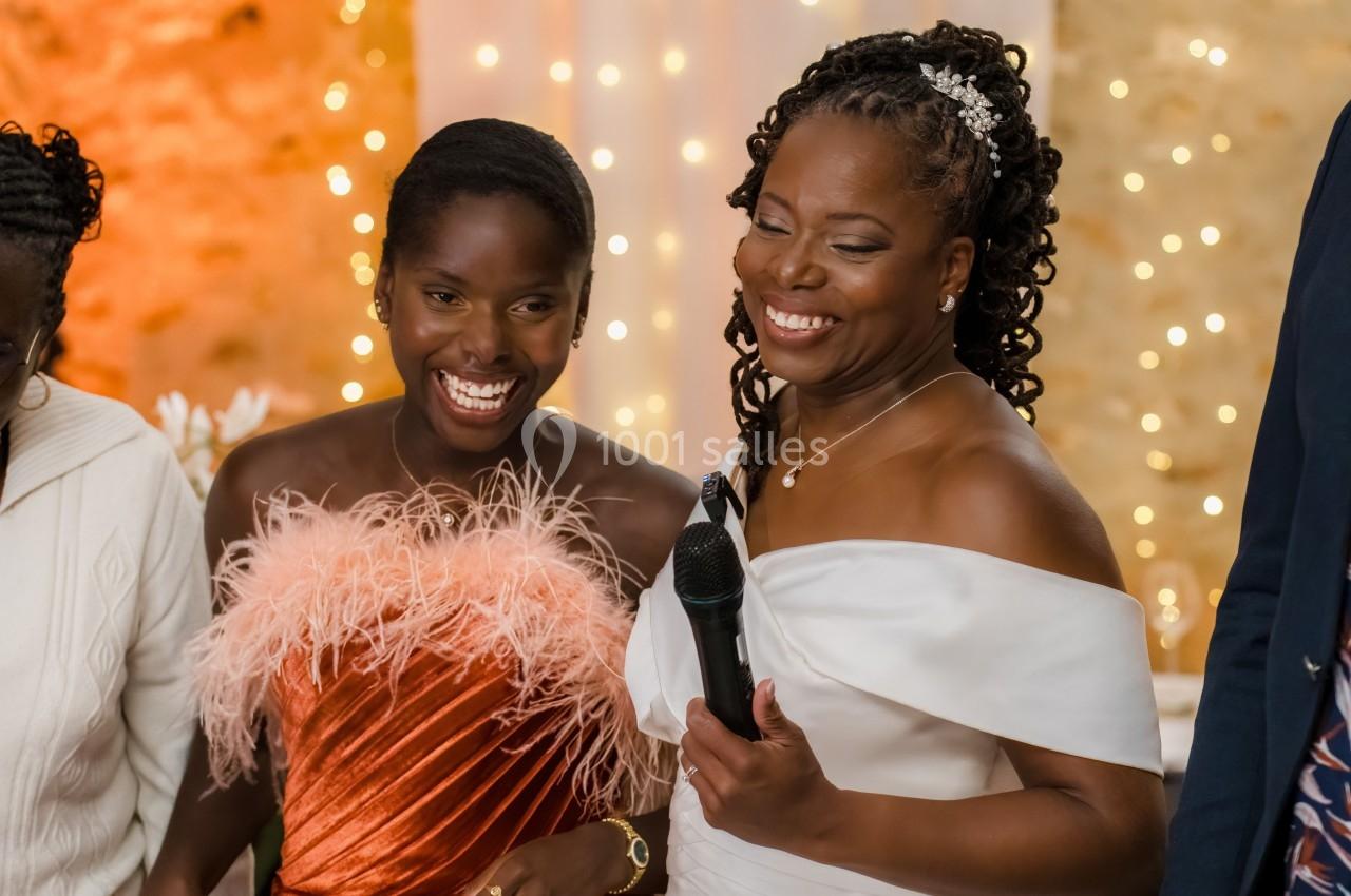 Deux femmes souriantes, l'une tenant un micro, dans un cadre festif avec des lumières en arrière-plan.