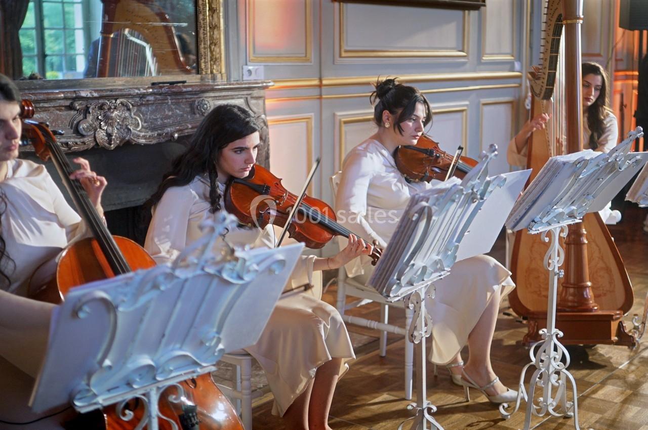 Quatre musiciennes jouent du violoncelle, du violon et de la harpe dans une salle élégante avec cheminée et moulures.