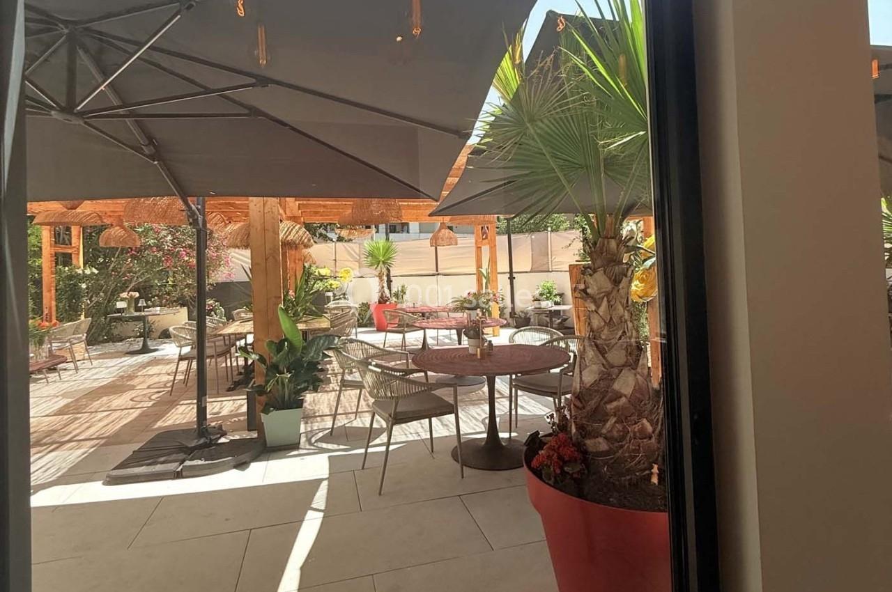 Terrasse ensoleillée avec tables, chaises, parasols et plantes, vue depuis une porte vitrée.