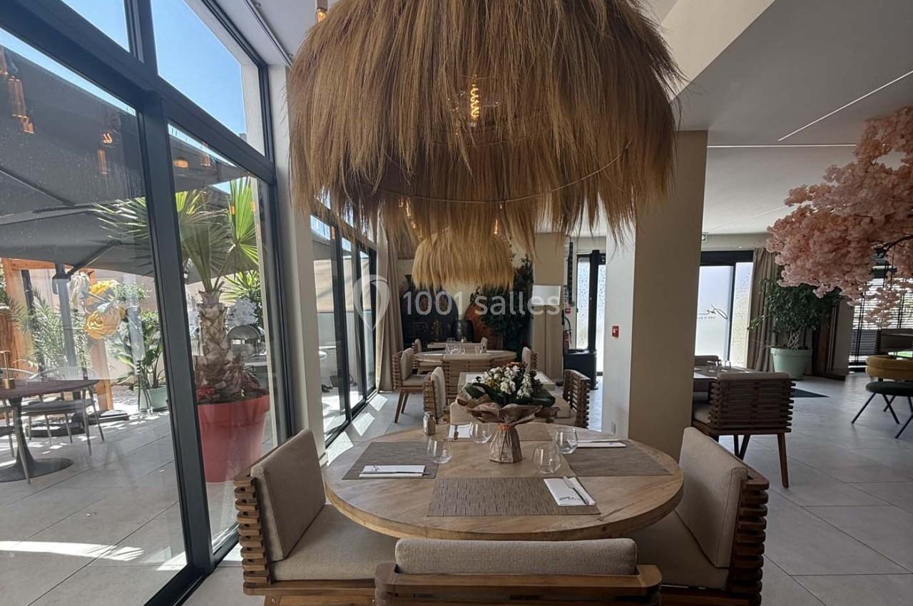 Salle de restaurant lumineuse avec grande baie vitrée, table ronde en bois et suspension en fibres naturelles.