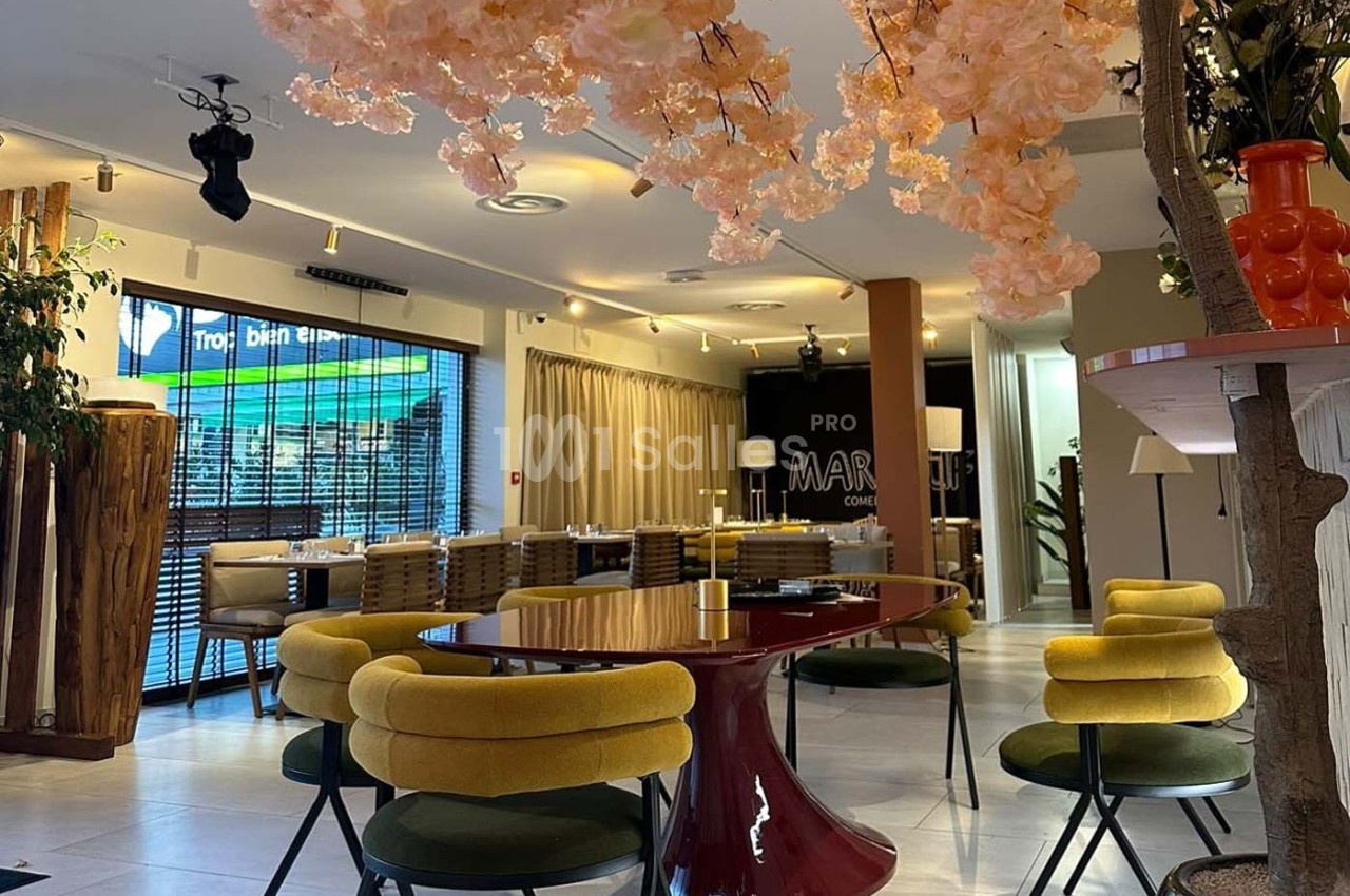 Salle de restaurant moderne avec mobilier coloré, table rouge, chaises jaunes et décoration florale suspendue.