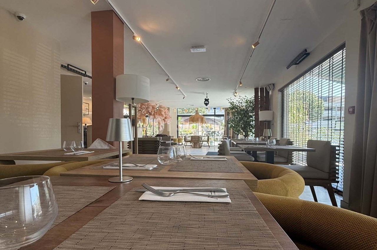 Salle de restaurant lumineuse avec tables dressées, chaises confortables et grandes baies vitrées donnant sur l'extérieur.