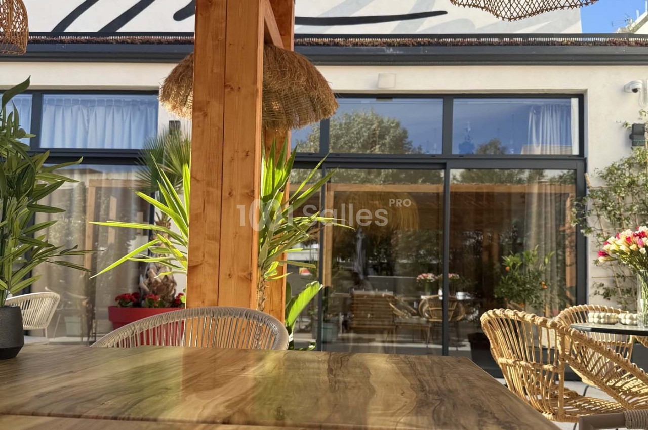 Terrasse ensoleillée avec table en bois, chaises en rotin et plantes, devant une baie vitrée donnant sur un intérieur…