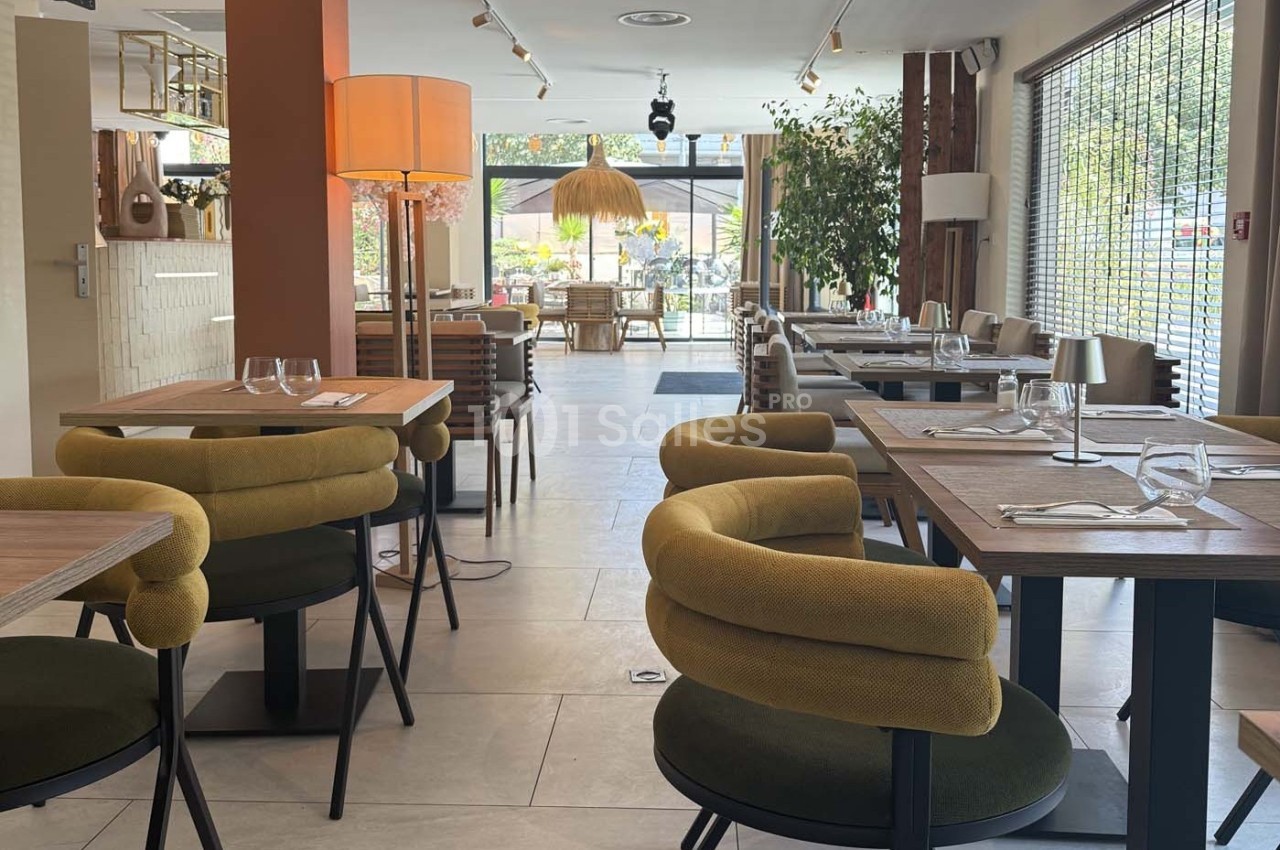 Salle de restaurant lumineuse avec tables en bois, chaises vertes et grandes baies vitrées donnant sur une terrasse.