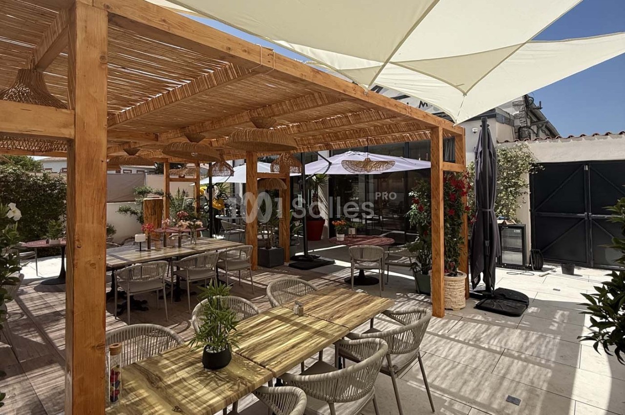 Terrasse extérieure avec pergola en bois, tables, chaises et plantes, ombragée par des voiles tendues.