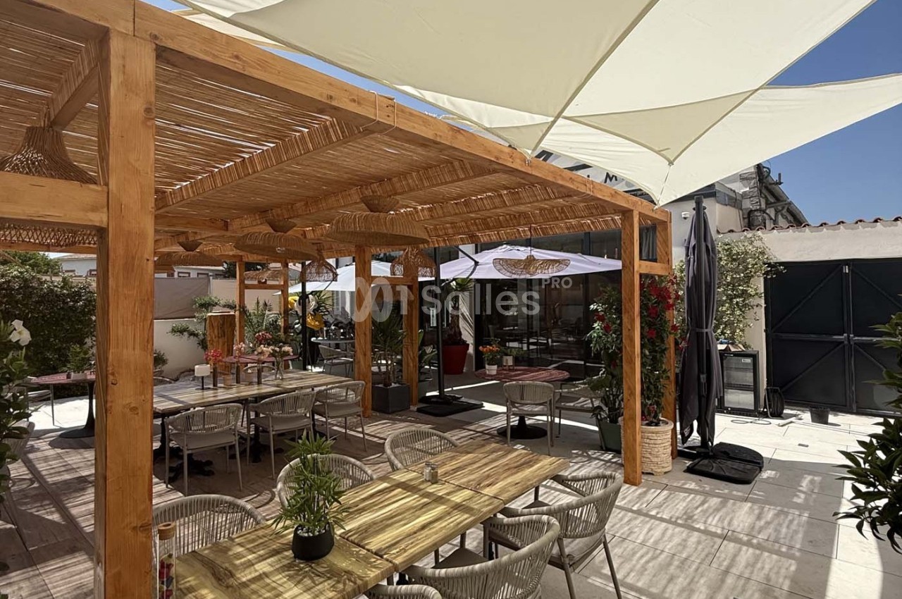 Terrasse extérieure avec pergola en bois, tables et chaises, entourée de plantes et protégée par des parasols blancs.