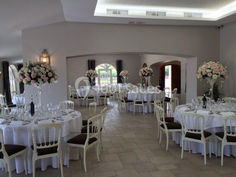 Salle de réception élégamment décorée avec des tables rondes, nappes blanches et bouquets de fleurs pastel.