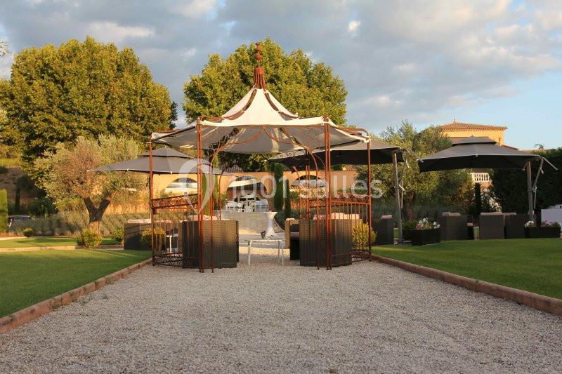 Allée en gravier menant à une pergola en fer forgé entourée de mobilier de jardin, avec pelouse et arbres en arrière-plan.