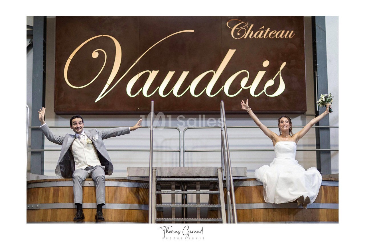 Un couple en tenue de mariage assis sur des barriques, souriant devant une enseigne ’Château Vaudois’.