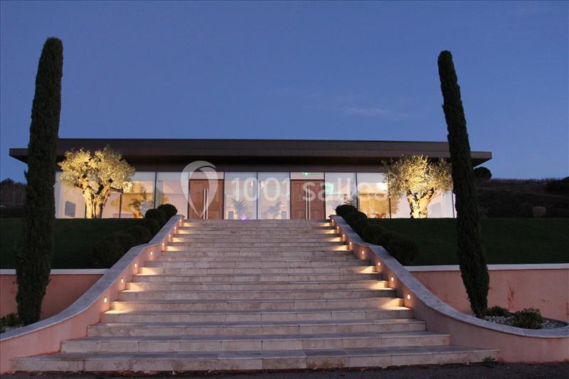 Escalier éclairé menant à un bâtiment moderne avec façade vitrée, entouré de cyprès et d'oliviers illuminés.