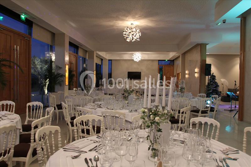 Salle de réception élégante avec tables rondes dressées, chaises blanches et chandeliers, éclairée par des lustres modernes.