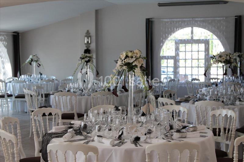 Salle de réception décorée avec des tables rondes, nappes blanches, chaises assorties et centres de table fleuris.