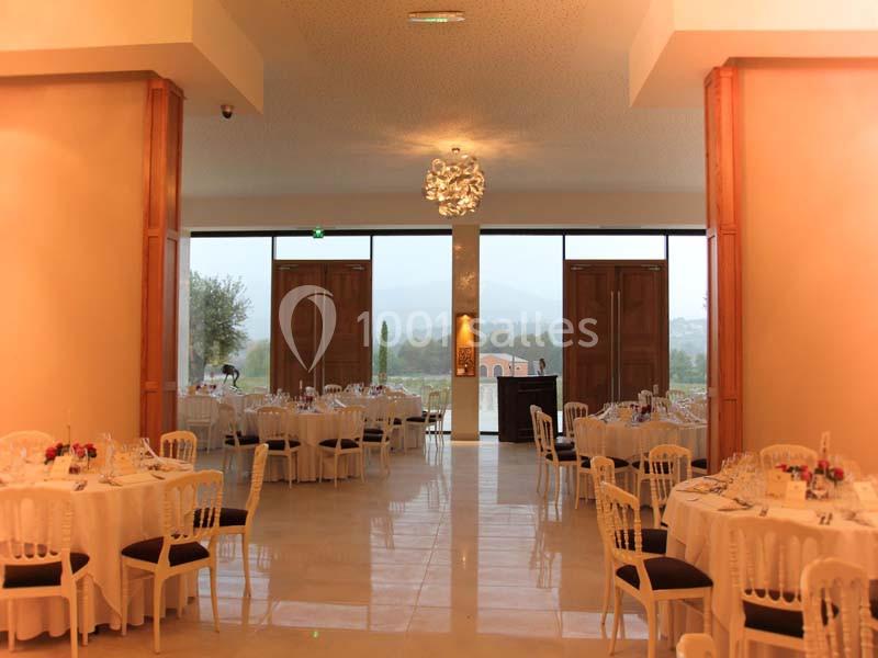 Salle de réception lumineuse avec tables rondes dressées, grandes baies vitrées et vue sur un paysage extérieur.