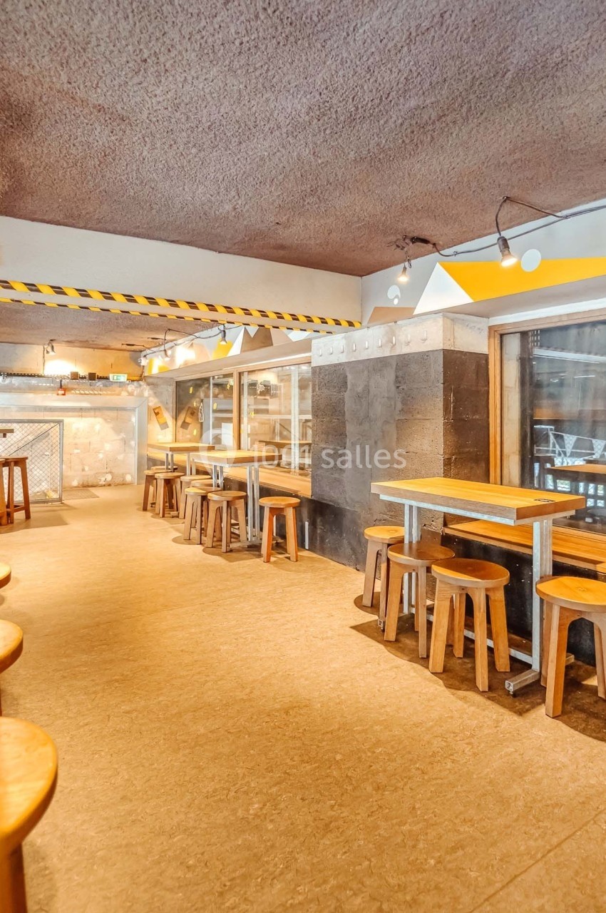 Salle de restaurant avec tables et tabourets en bois, éclairage suspendu et murs en béton brut.