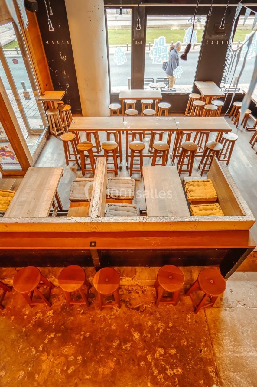 Vue plongeante d'un café avec tables en bois, tabourets alignés et grandes fenêtres donnant sur l'extérieur.
