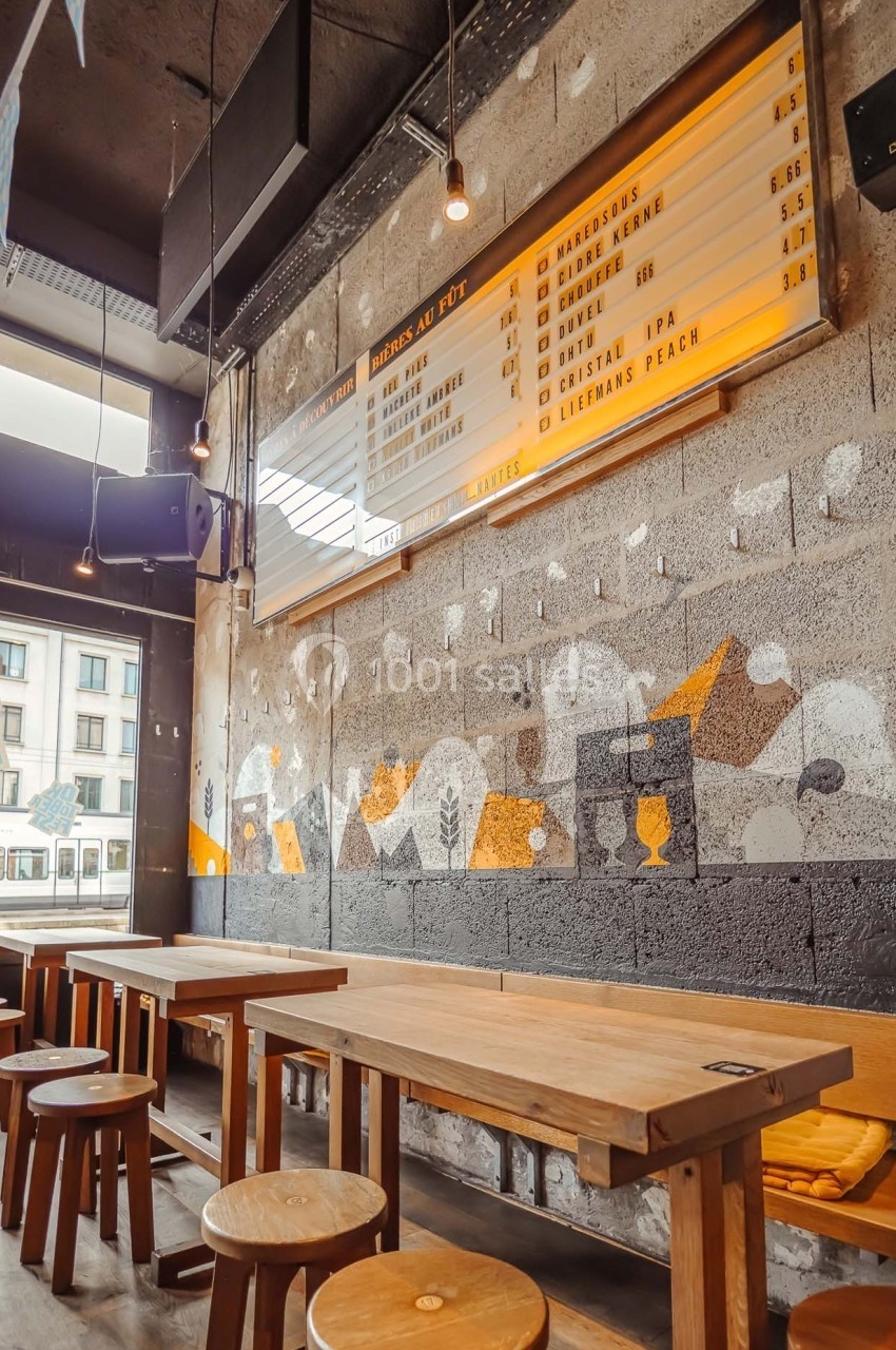 Intérieur d'un bar avec tables en bois, tabourets, mur décoré de fresques et menu affiché sur un panneau lumineux.