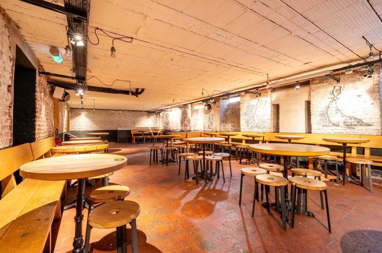 Salle avec tables et tabourets en bois, murs en briques apparentes et éclairage suspendu dans une ambiance industrielle.