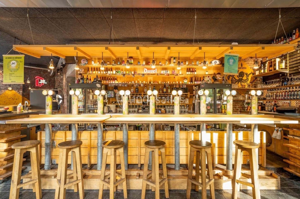 Bar en bois avec tabourets alignés, comptoir équipé de tireuses à bière et étagères remplies de bouteilles en arrière-plan.