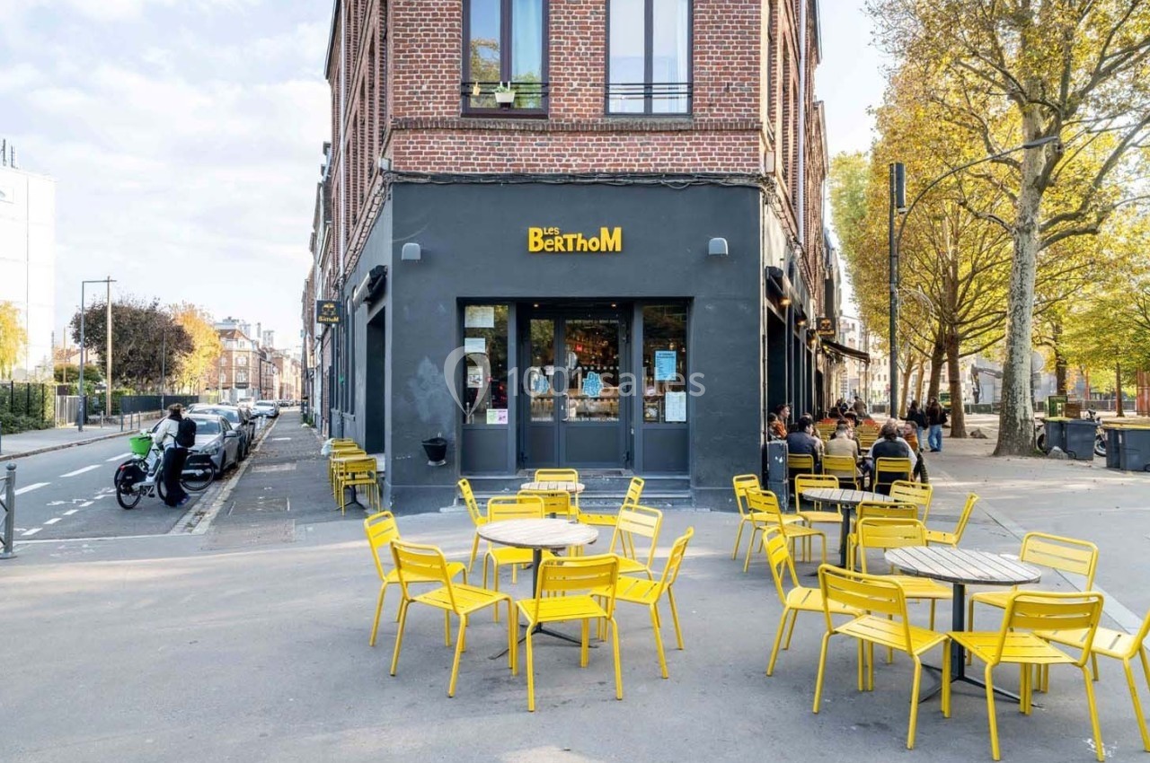 Terrasse avec des tables et chaises jaunes devant un bar en briques nommé ’Les Berthom’, situé à un carrefour urbain.