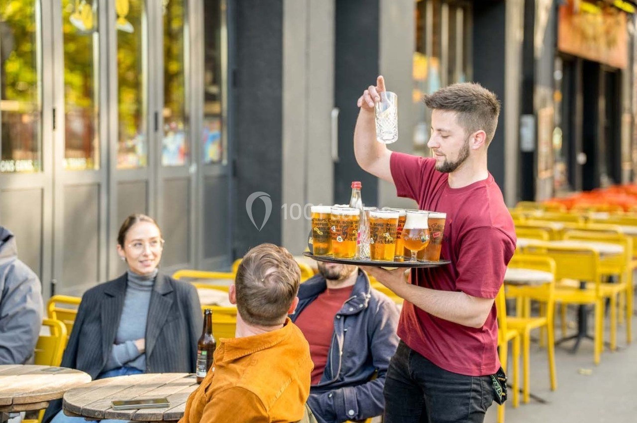 Un serveur apporte un plateau de bières et un verre d'eau à des clients assis en terrasse.