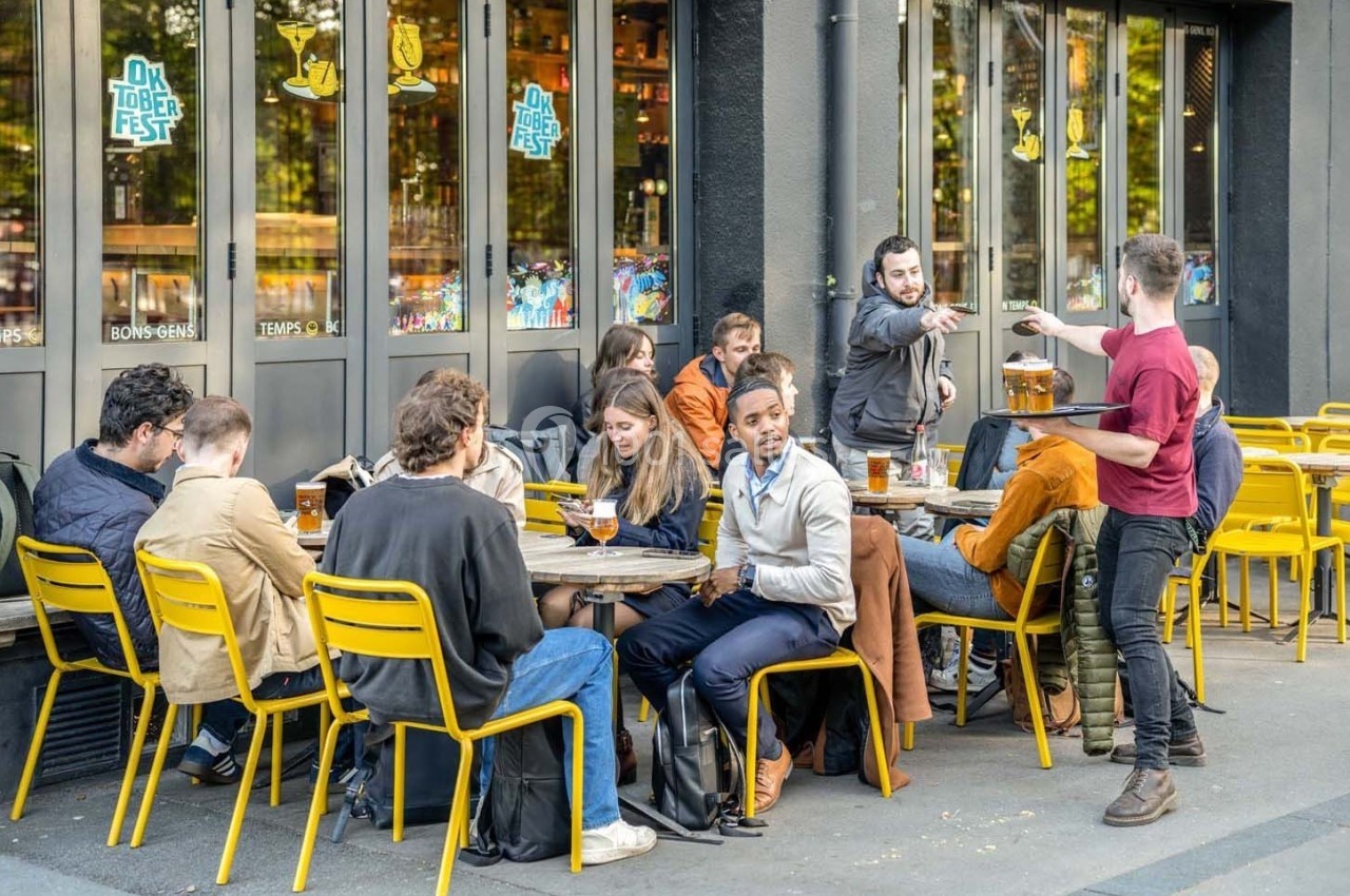 Des personnes assises en terrasse d'un café, discutant et partageant des boissons, avec un serveur apportant des verres.