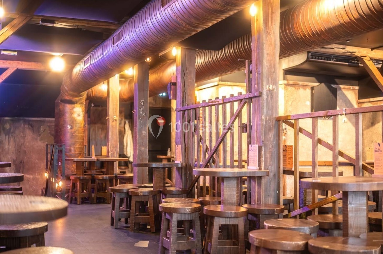 Salle de bar avec des tables et tabourets en bois, éclairage chaleureux et conduits métalliques apparents au plafond.