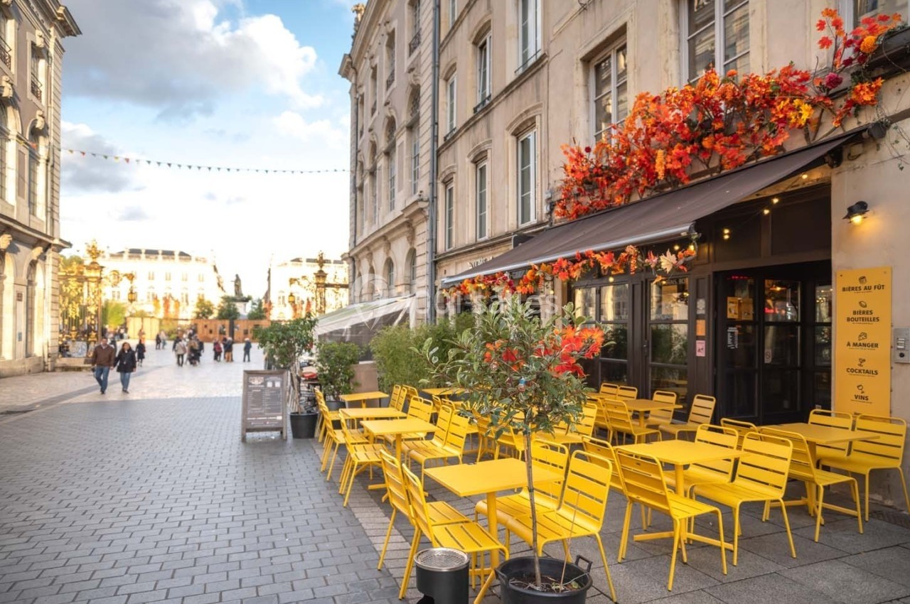 Terrasse de café avec chaises jaunes vives, située sur une place pavée bordée de bâtiments anciens et décorée de fleurs.