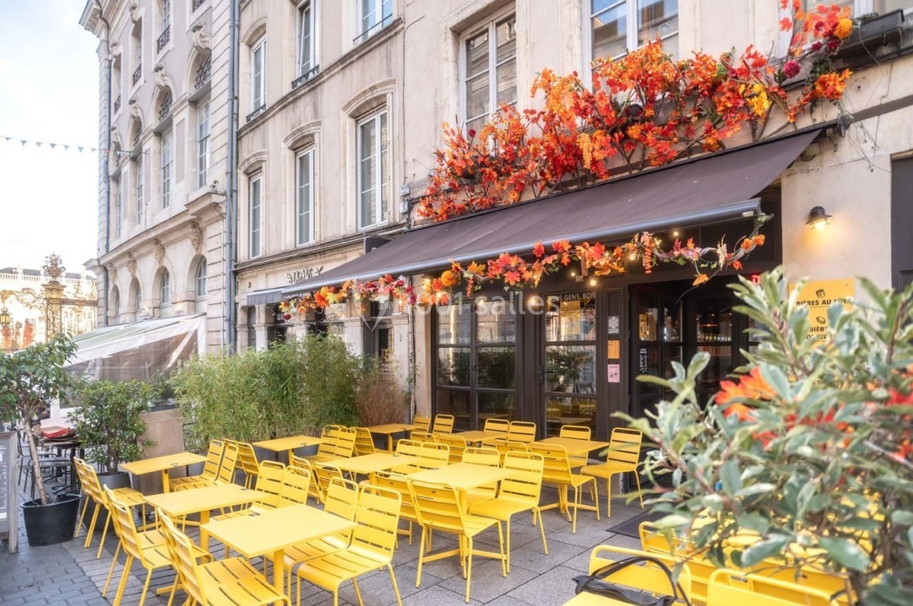 Terrasse d'un café avec des tables et chaises jaunes, façade décorée de feuillage automnal, dans une rue bordée d'immeubles.