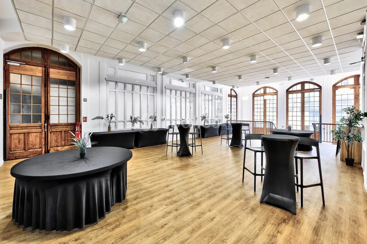 Salle lumineuse avec parquet, grandes fenêtres cintrées, tables rondes et hautes noires, et plantes décoratives.