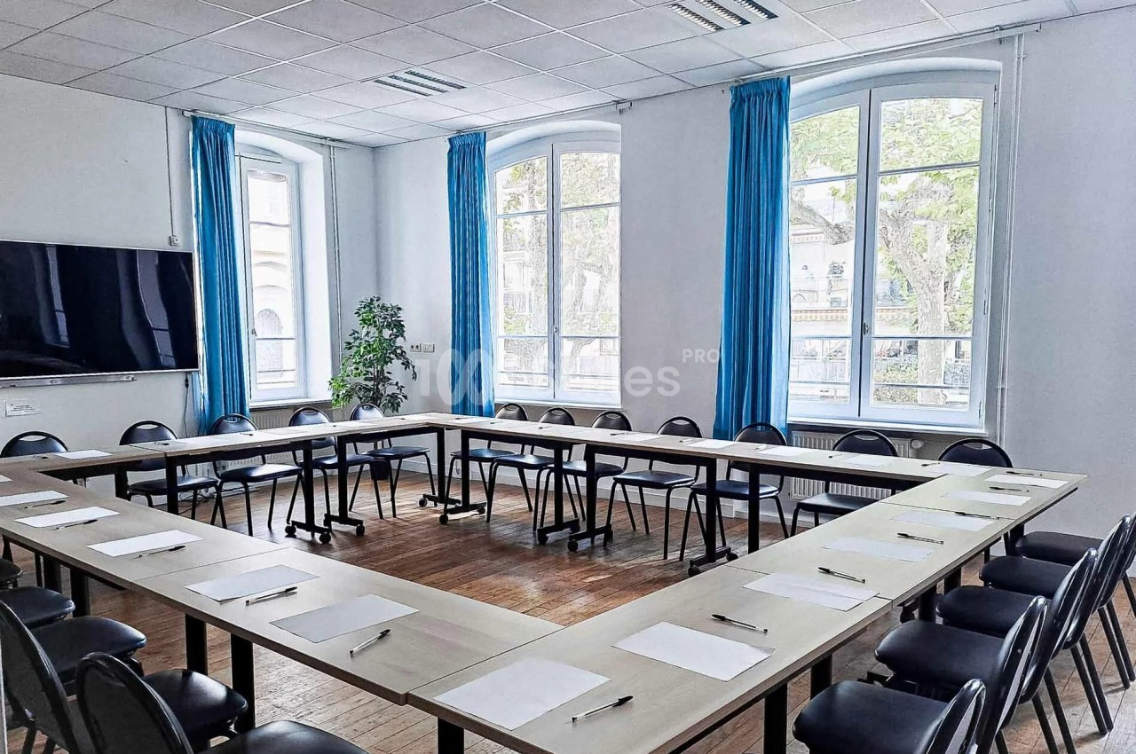 Salle de réunion lumineuse avec tables en U, chaises noires, grandes fenêtres et rideaux bleus.