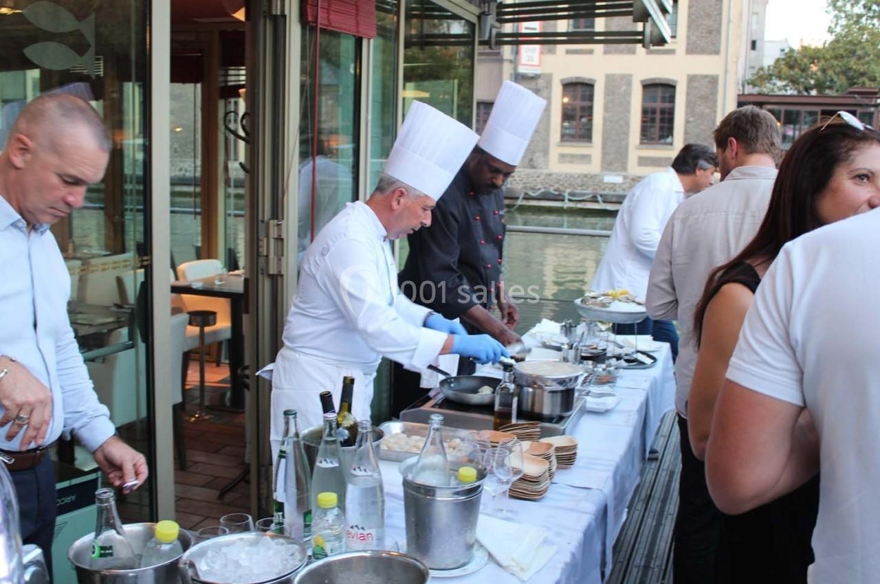 Des chefs en tenue blanche et noire préparent des plats sur une table en extérieur près d'un canal.