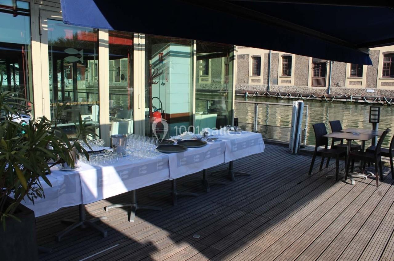 Terrasse en bois avec une table dressée pour un buffet, située au bord de l'eau près d'un bâtiment en briques.