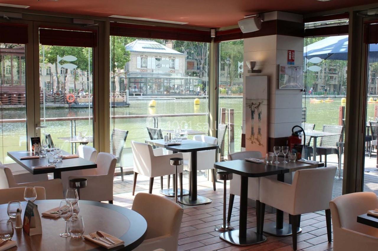 Salle de restaurant lumineuse avec vue sur un canal, tables dressées et grandes baies vitrées donnant sur l'extérieur.