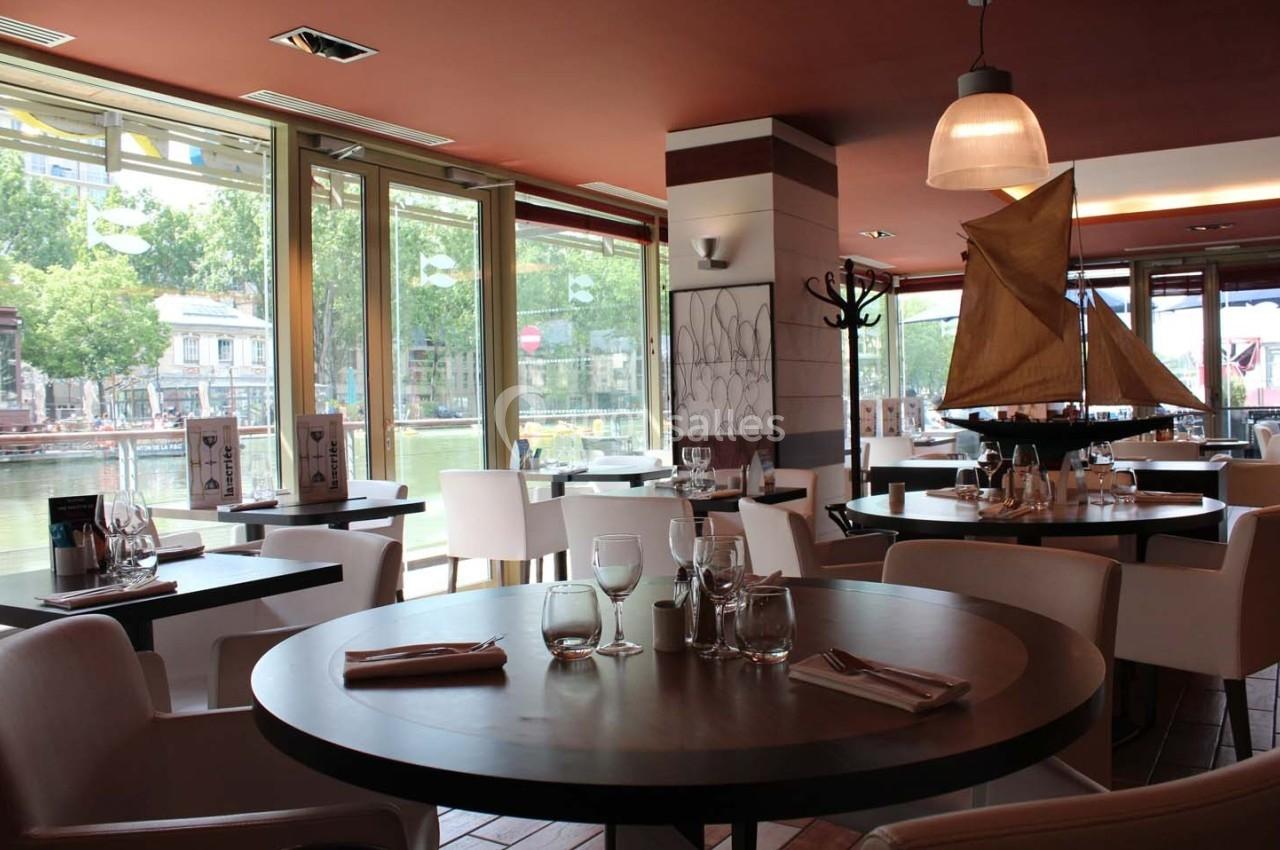 Salle de restaurant lumineuse avec tables dressées, vue sur un canal à travers de grandes baies vitrées.