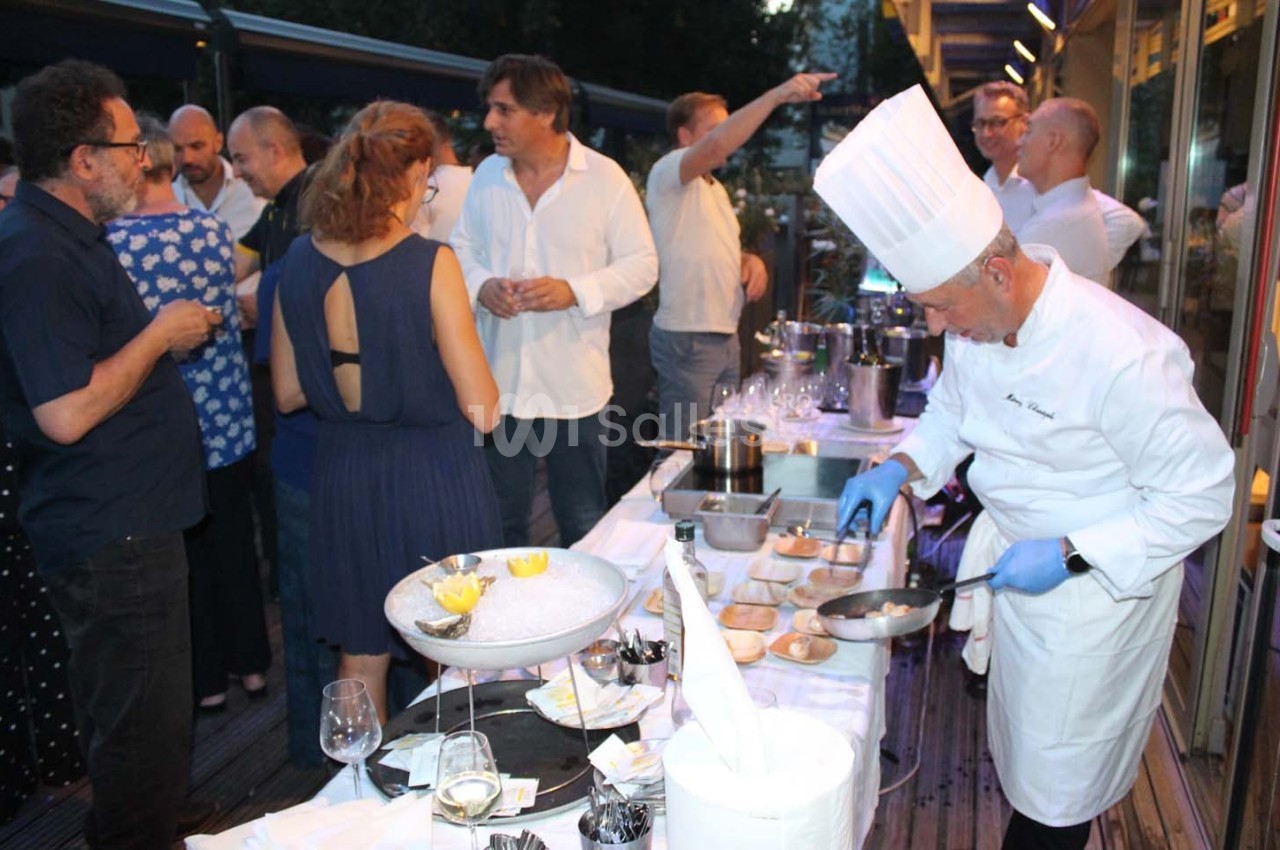 Un chef prépare des plats devant des convives rassemblés autour d'une table lors d'un événement en extérieur.