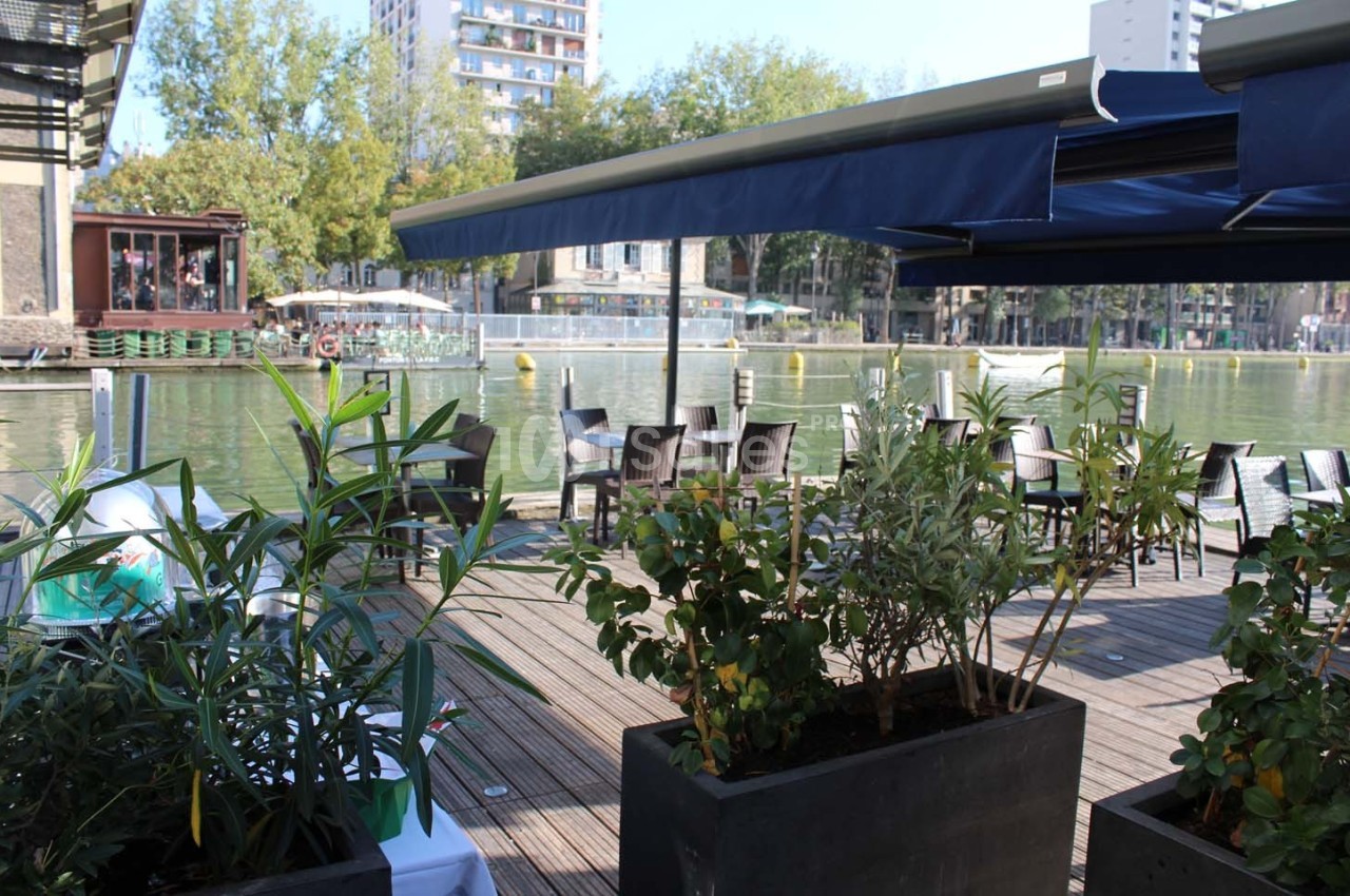 Terrasse en bois au bord d'un canal, avec des plantes en pot, des tables et des chaises sous un auvent bleu.