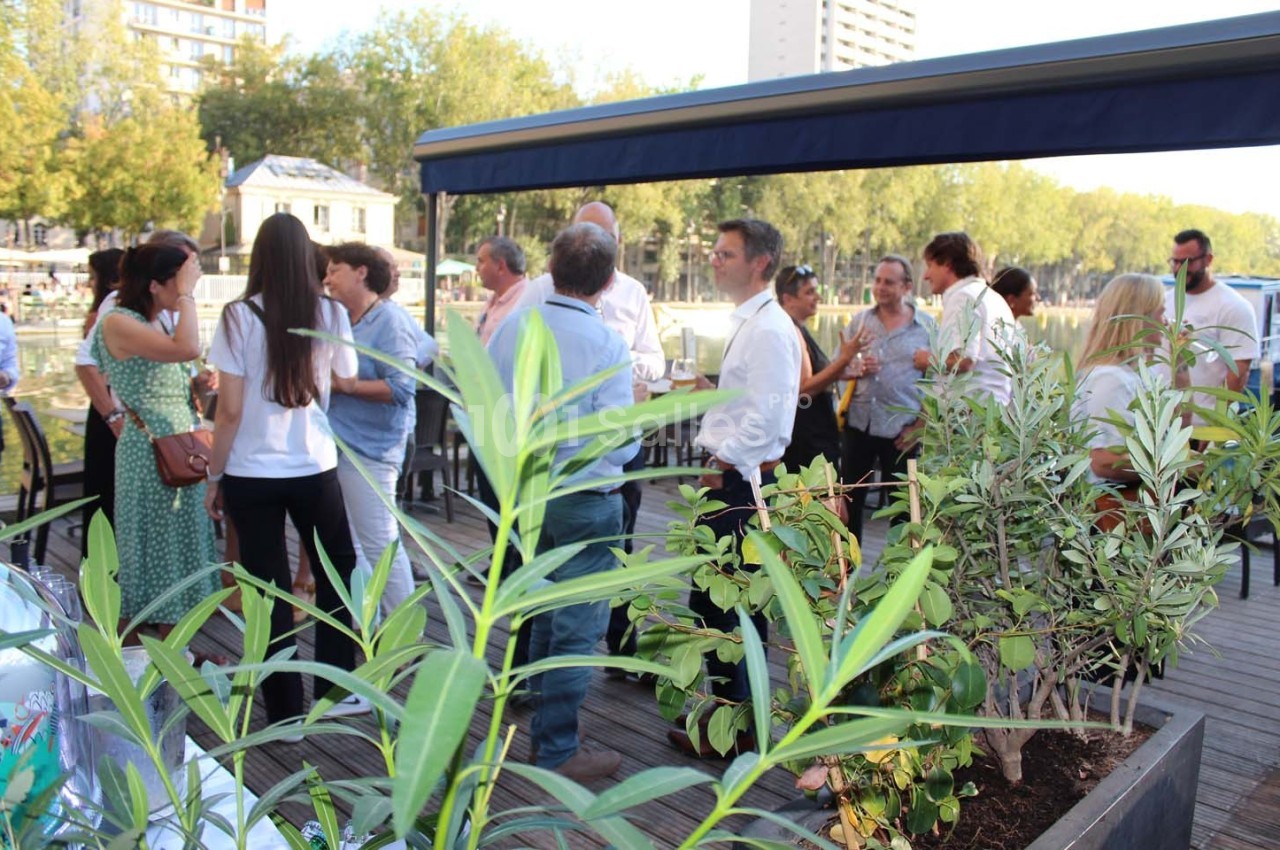 Groupe de personnes discutant sur une terrasse en bois bordée de plantes, avec des arbres et des bâtiments en arrière-plan.