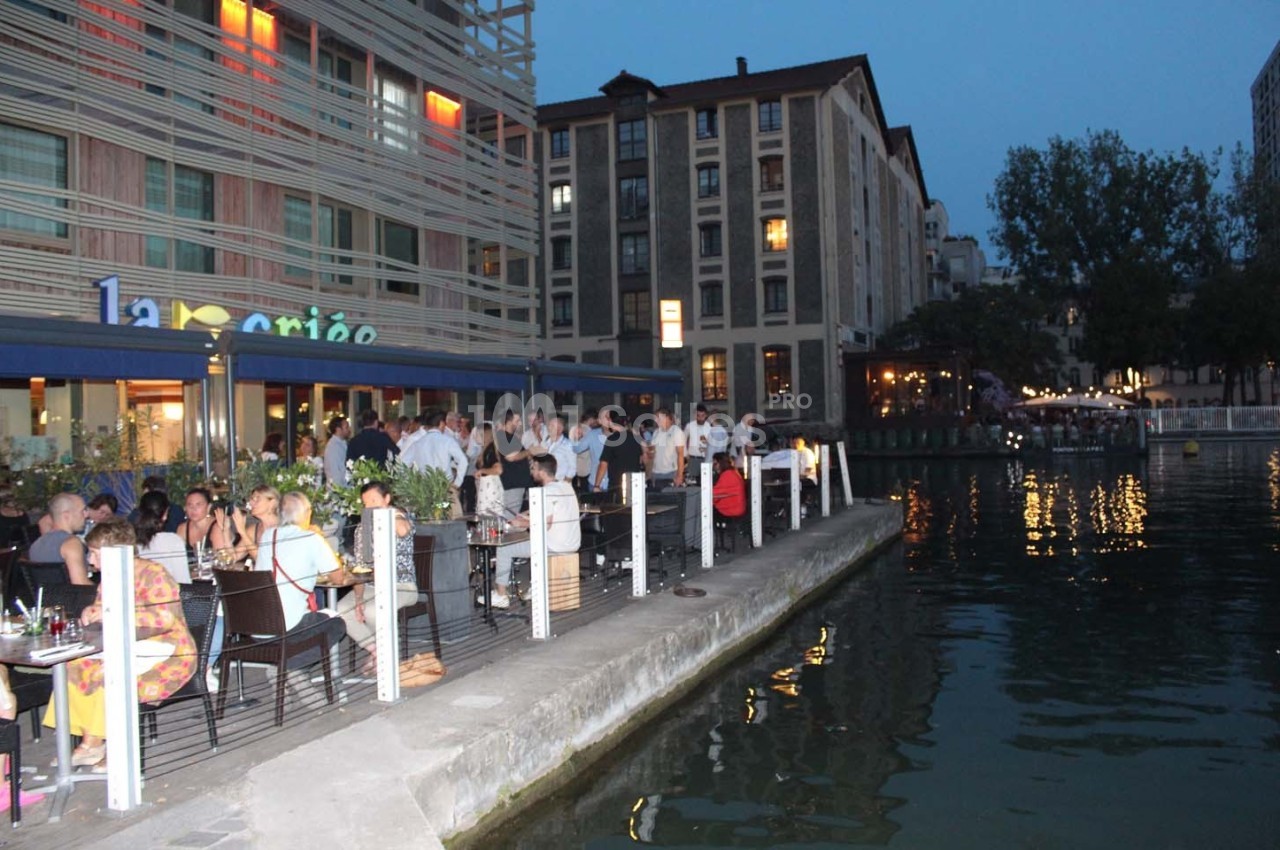 Des personnes dînent en terrasse au bord d'un canal, près d'un restaurant éclairé en soirée.