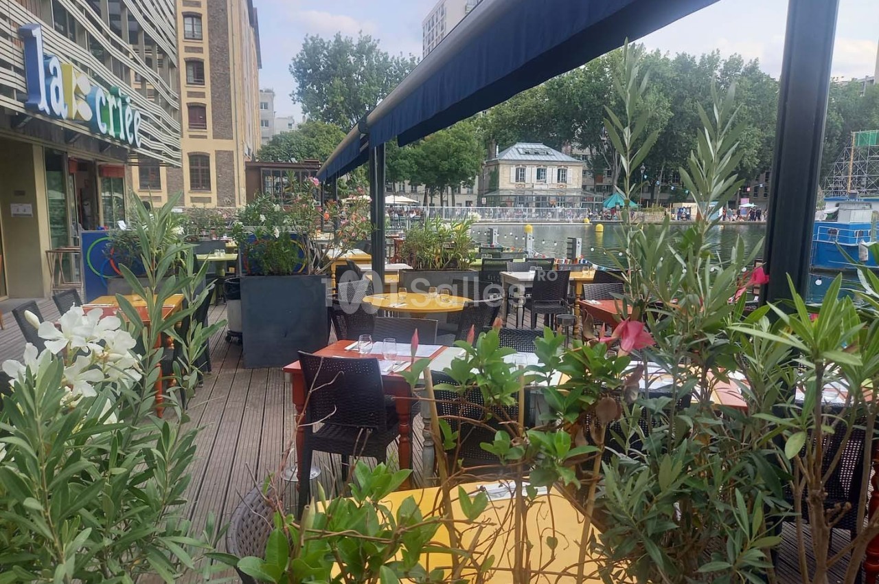 Terrasse de restaurant avec tables dressées, entourée de plantes, donnant sur un canal et des bâtiments en arrière-plan.