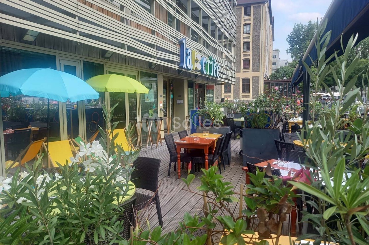 Terrasse d'un café avec tables, chaises et parasols colorés, entourée de plantes, devant un immeuble moderne.