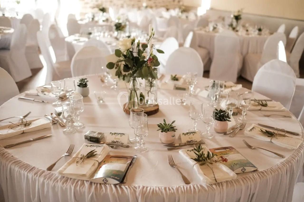 Table ronde décorée pour un événement, avec nappes blanches, fleurs, menus et couverts disposés élégamment.