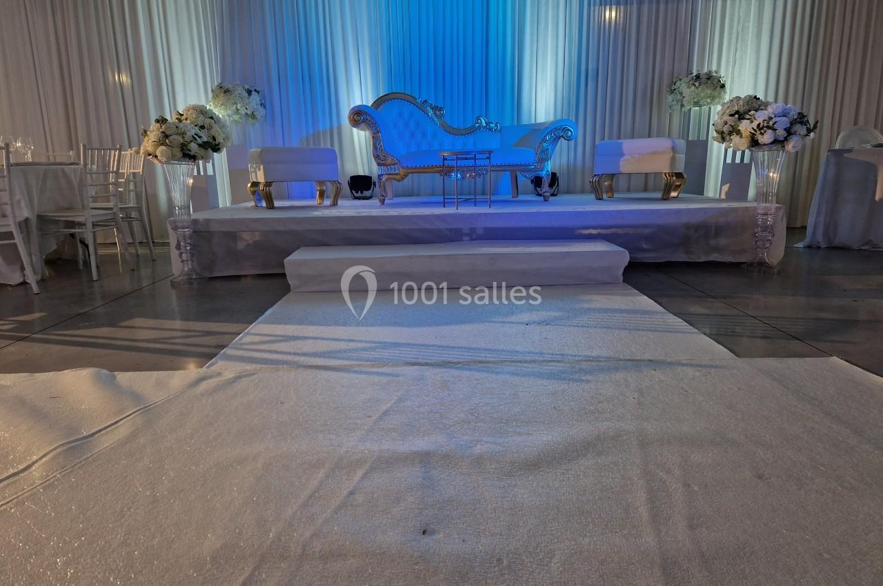 Scène de mariage avec canapé élégant, chaises blanches, fleurs et éclairage bleu sur fond de rideaux blancs.