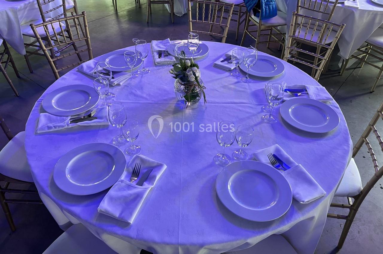 Table ronde dressée avec nappes blanches, assiettes, couverts, verres et un centre de table végétal, entourée de chaises.